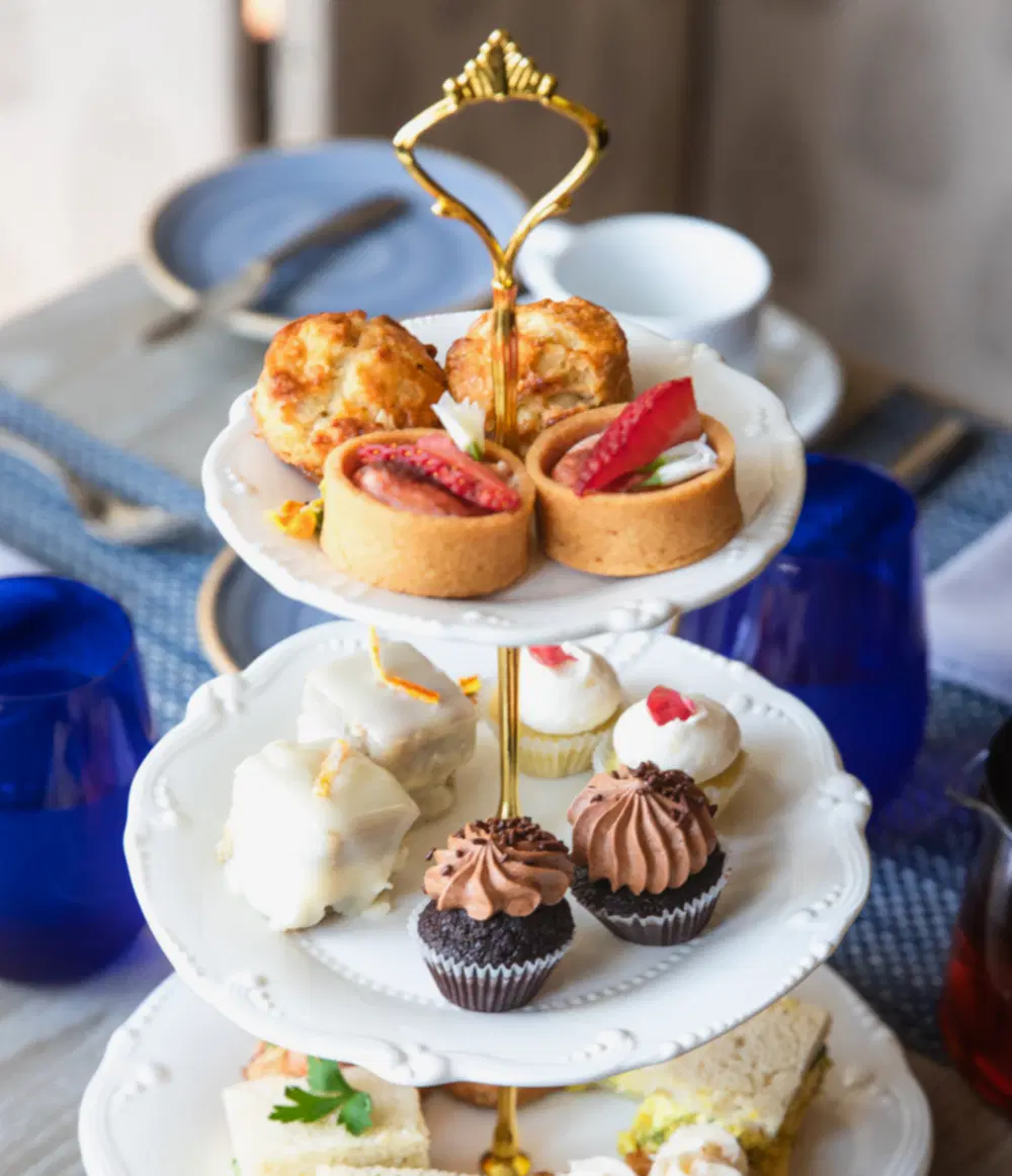 Three-tiered tray with assorted pastries, cupcakes, tarts, scones, and finger sandwiches, set on a table with blue glasses and plates in the background.