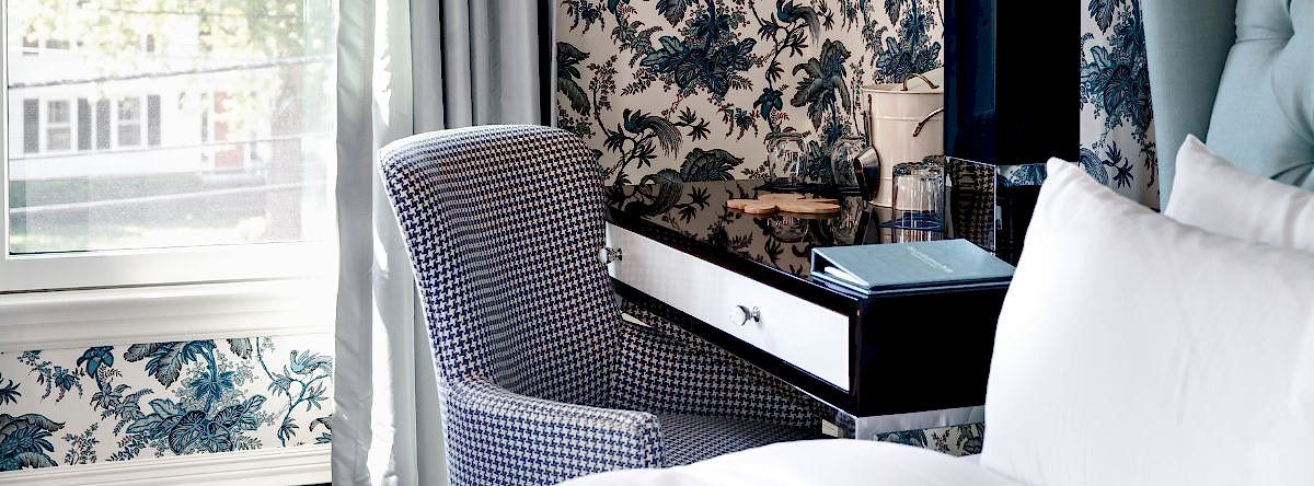 Patterned chair at a black desk with a book, lamp, and glasses beside a bed in a room with floral wallpaper and a window with sheer curtains.