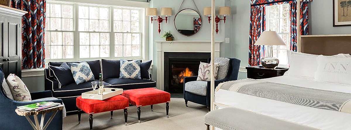 Cozy bedroom with a blue sofa, red ottomans, fireplace, large bed, and patterned curtains. Sunlight streams through two large windows.