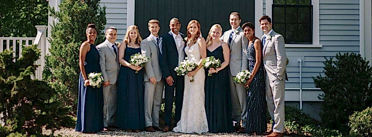 A wedding party poses outside; the bride is in white, the groom in a suit, with bridesmaids in navy dresses and groomsmen in gray suits.