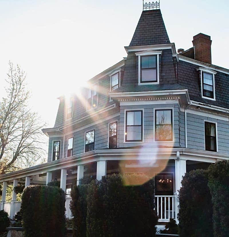 A large, gray Victorian-style house with a porch, black roof, and sunlight streaming over the roof, surrounded by bushes and trees.