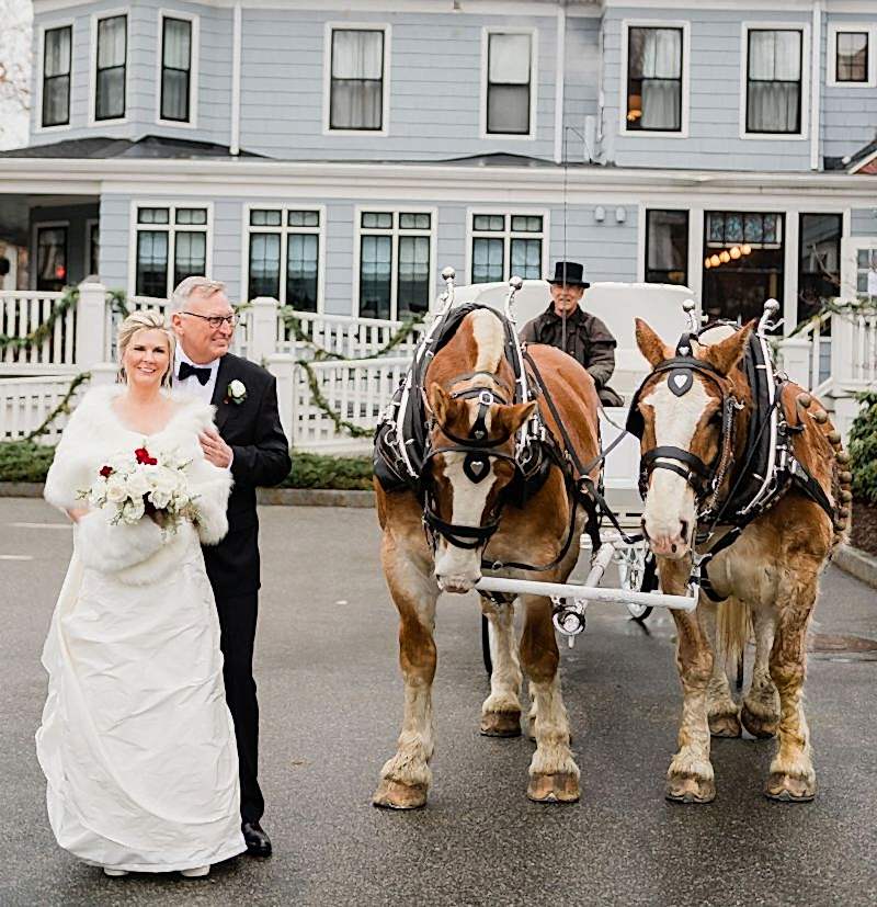 A smiling bride and groom stand in front of a white house with a horse-drawn carriage and two brown horses.