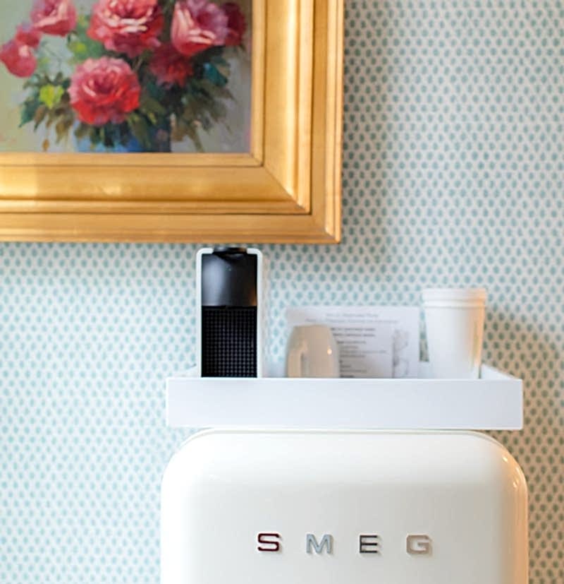 A white SMEG fridge with items on top, beneath a gold-framed painting of red and pink flowers on a blue patterned wall.