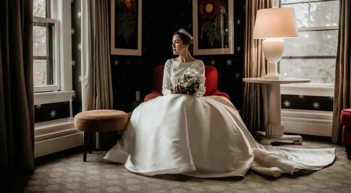 A bride in a white gown sits on a red chair, holding a bouquet, by a window in a dimly lit room with floral decor and a lit table lamp.