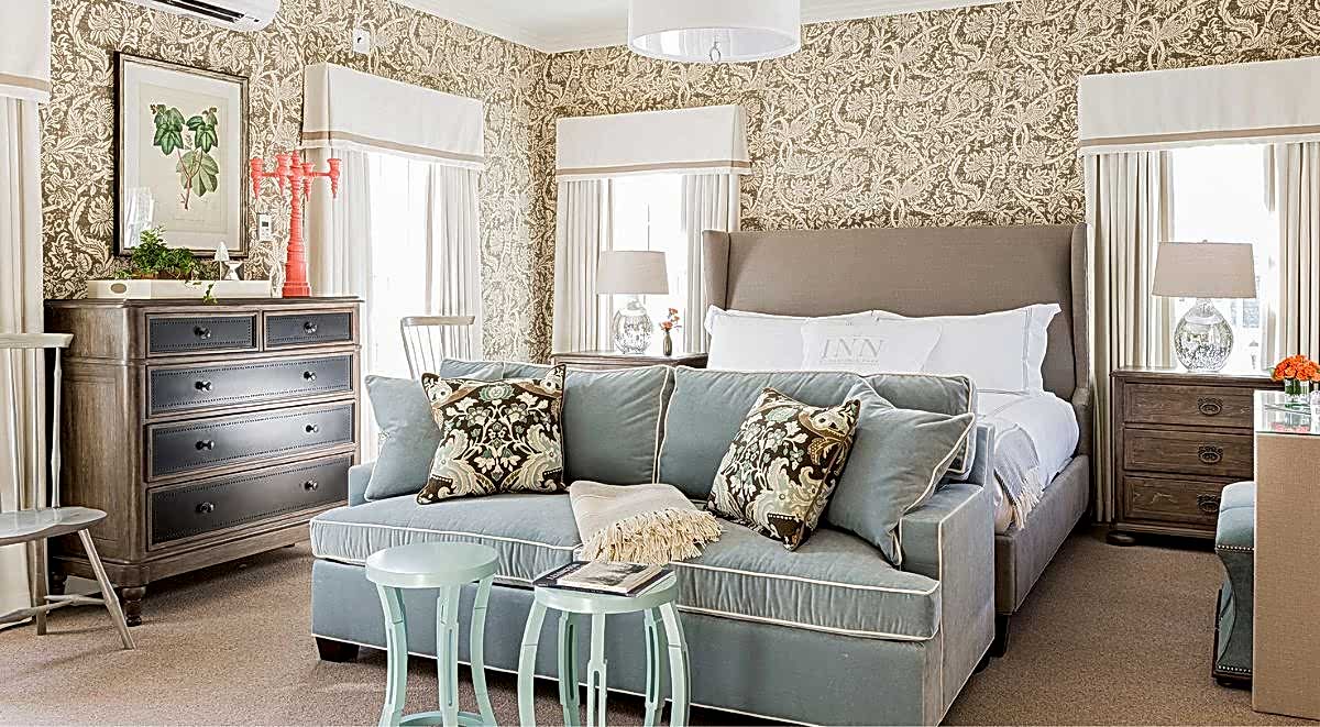 Elegant bedroom with a patterned wallpaper, a bed with white linens, a blue loveseat, and wood furniture, softly lit by natural light from windows.