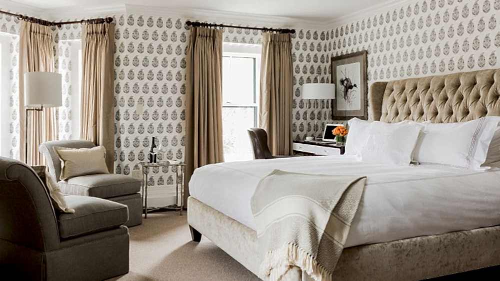 Bright, elegant bedroom with a large bed, beige tufted headboard, light patterned wallpaper, armchair, desk, and windows with tan curtains.