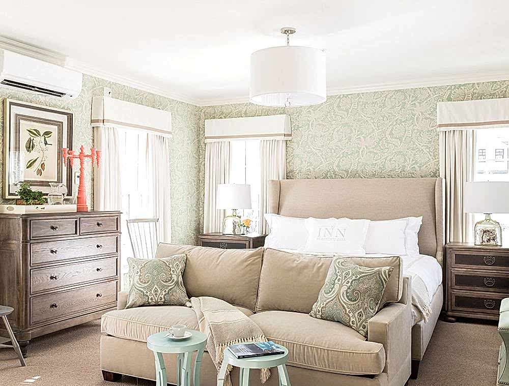 Bright, elegant bedroom with a beige bed, light sofa, patterned pillows, wooden dressers, and green floral wallpaper, filled with natural light.