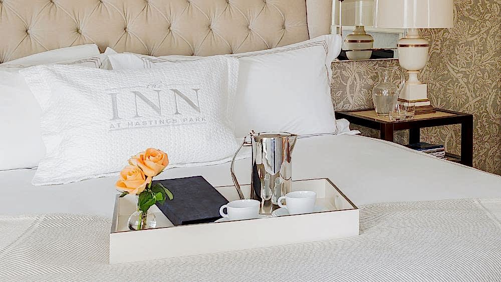A hotel bed with white linens, a pillow labeled Inn at Hastings Park, and a tray holding two cups, a silver pot, and a vase with orange roses.