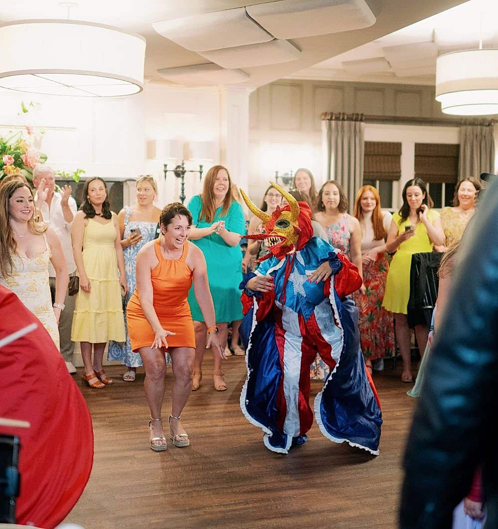 A person in a colorful traditional mask dances while a group of women in bright dresses cheer and watch in a festive indoor setting.