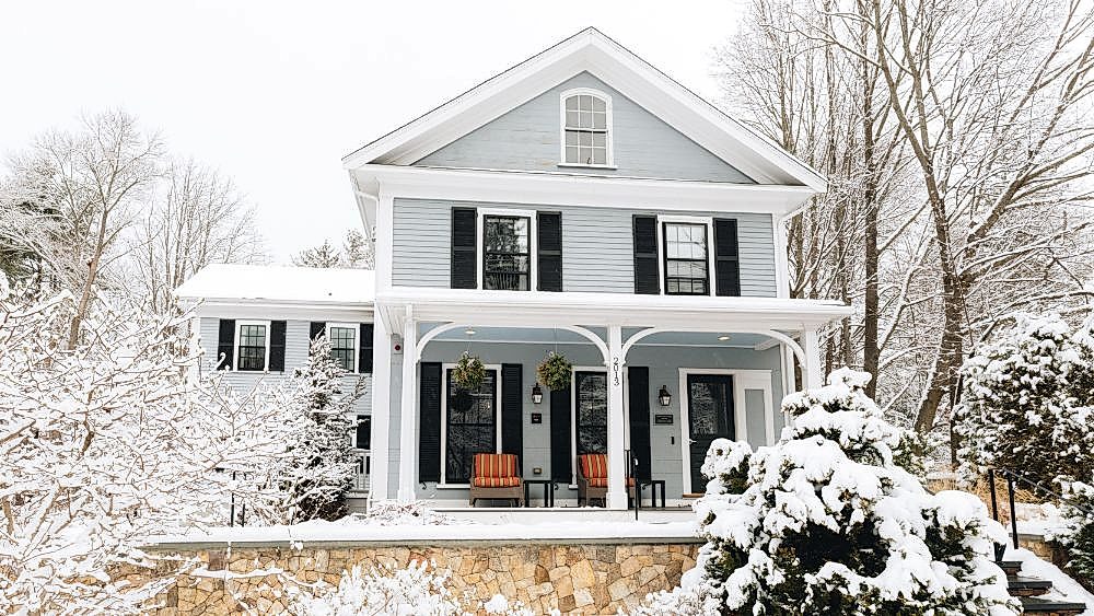 Light blue house with black shutters, white trim, and two orange chairs on the porch, surrounded by snow-covered trees and bushes in winter.