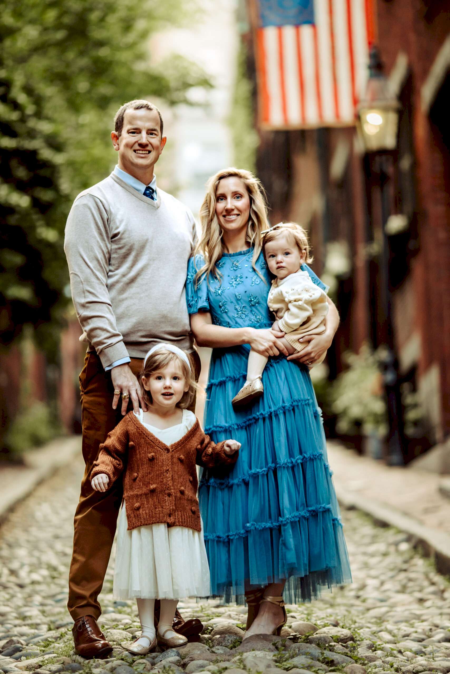 A family of four poses together on a cobblestone street, with greenery and an American flag in the background.