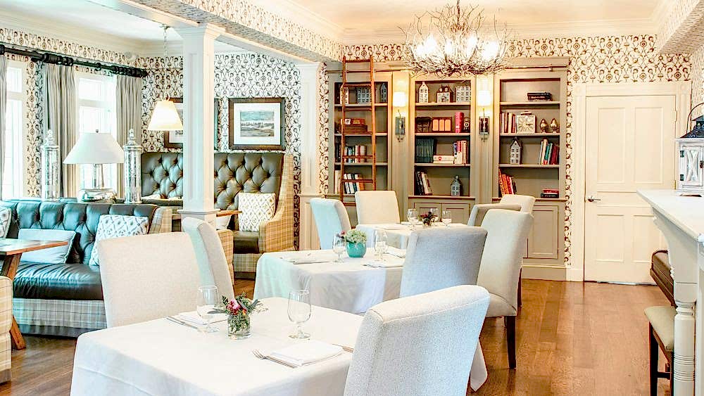 Elegant dining room with white tablecloths, upholstered chairs, bookshelves, and large windows letting in natural light; floral decor on tables.