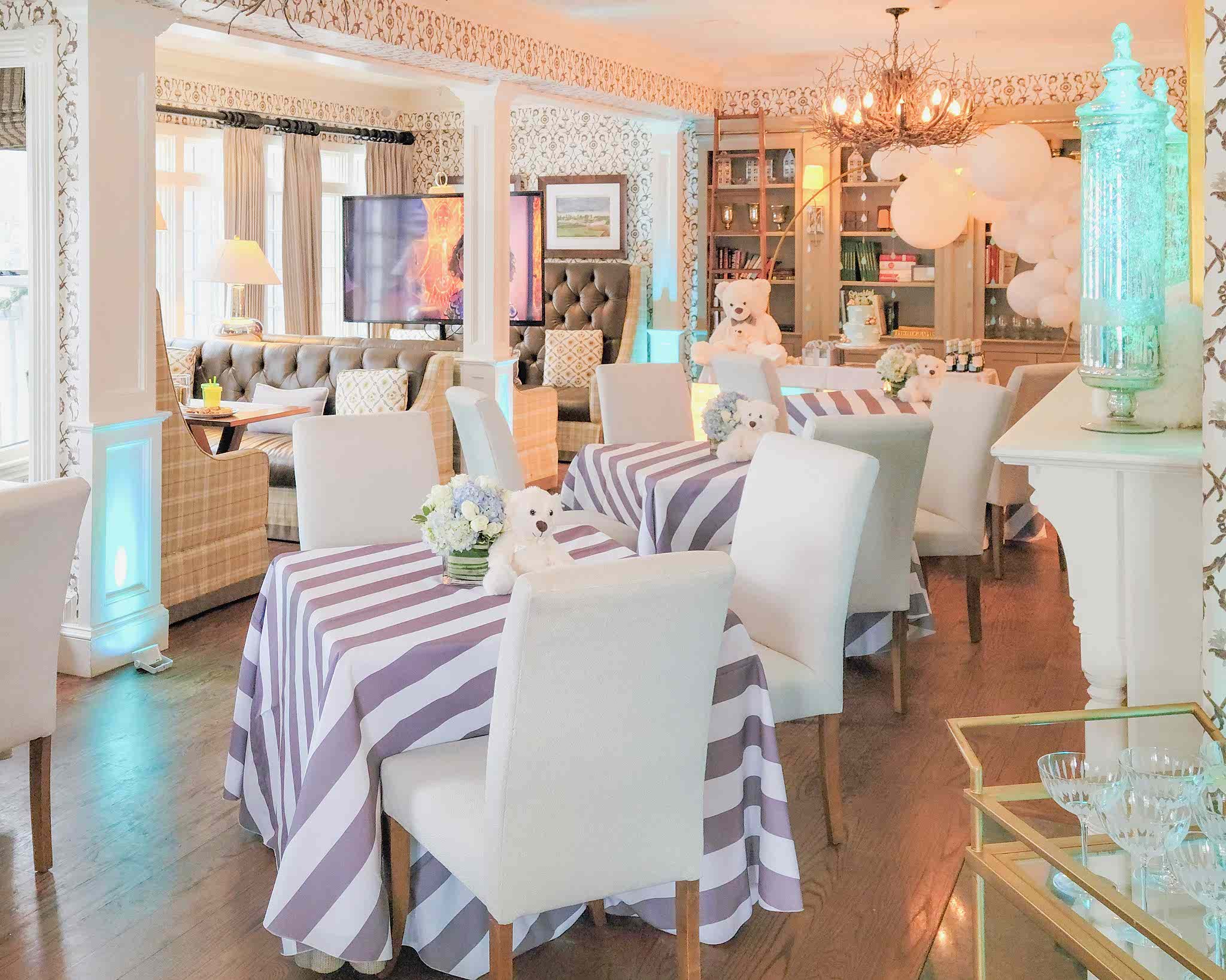 Elegant room with tables draped in purple and white striped cloths, white chairs, floral centerpieces, and festive decorations including balloons and a teddy bear.