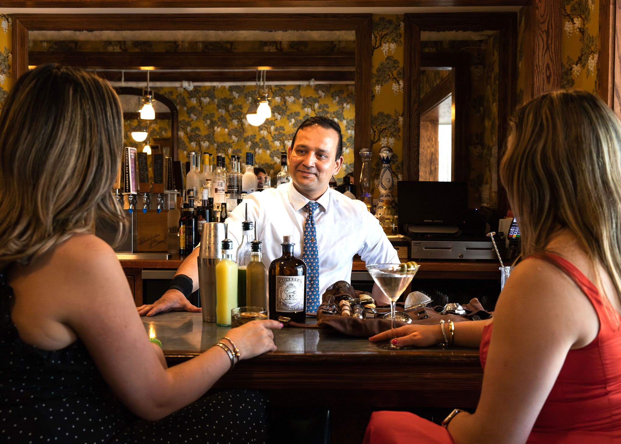 A bartender in a white shirt and tie smiles while talking to two women seated at a bar with drinks and cocktail ingredients in front of them.