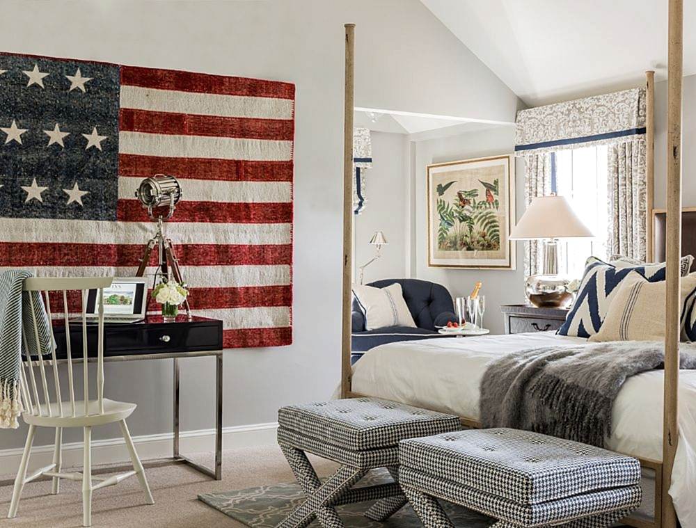 Bedroom with a four-poster bed, American flag wall art, desk with chair, patterned ottomans, and large window with drapes letting in natural light.