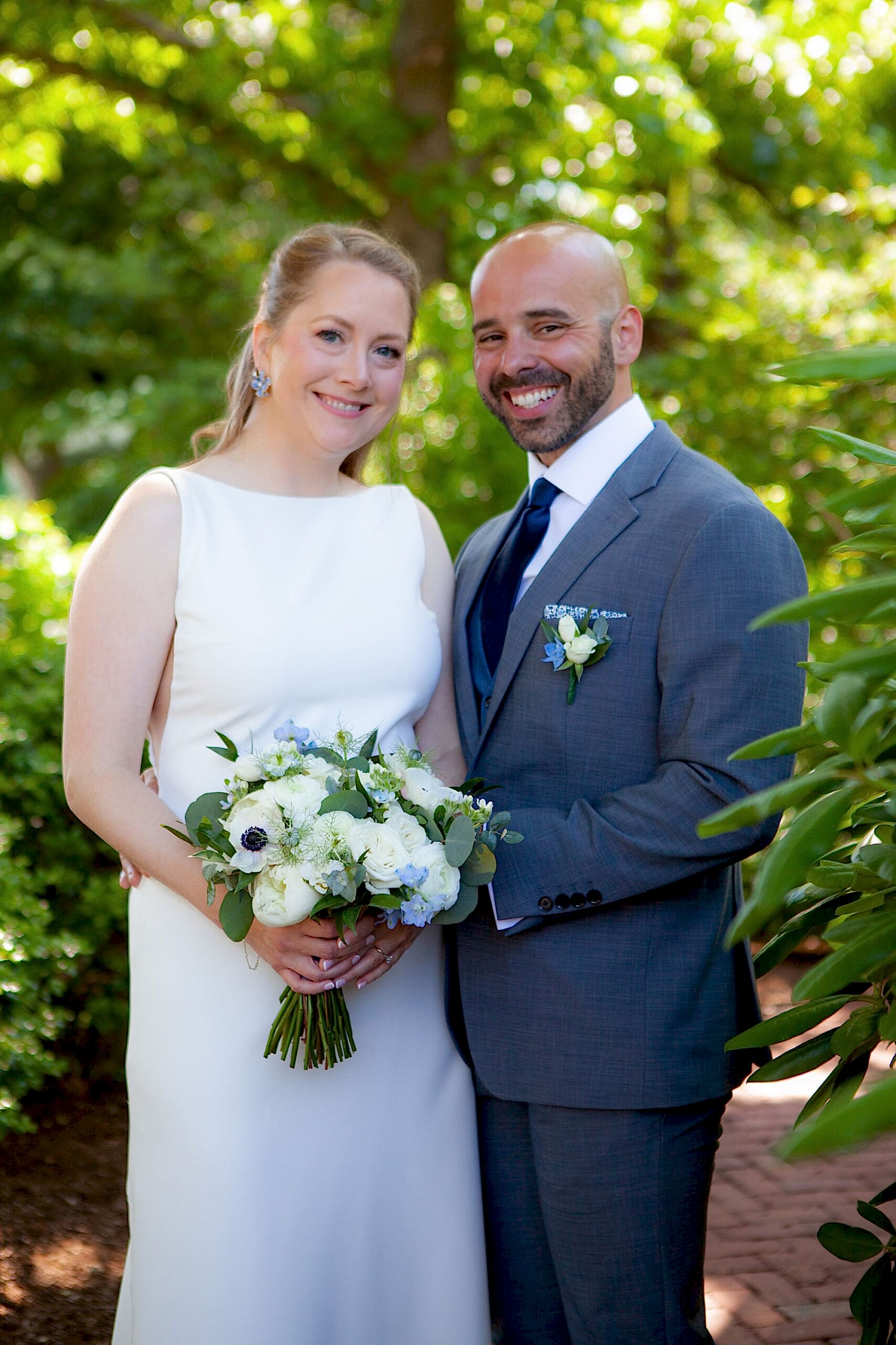 A bride in a white dress and a groom in a gray suit smile together outdoors, surrounded by greenery, holding a bouquet of white and blue flowers.