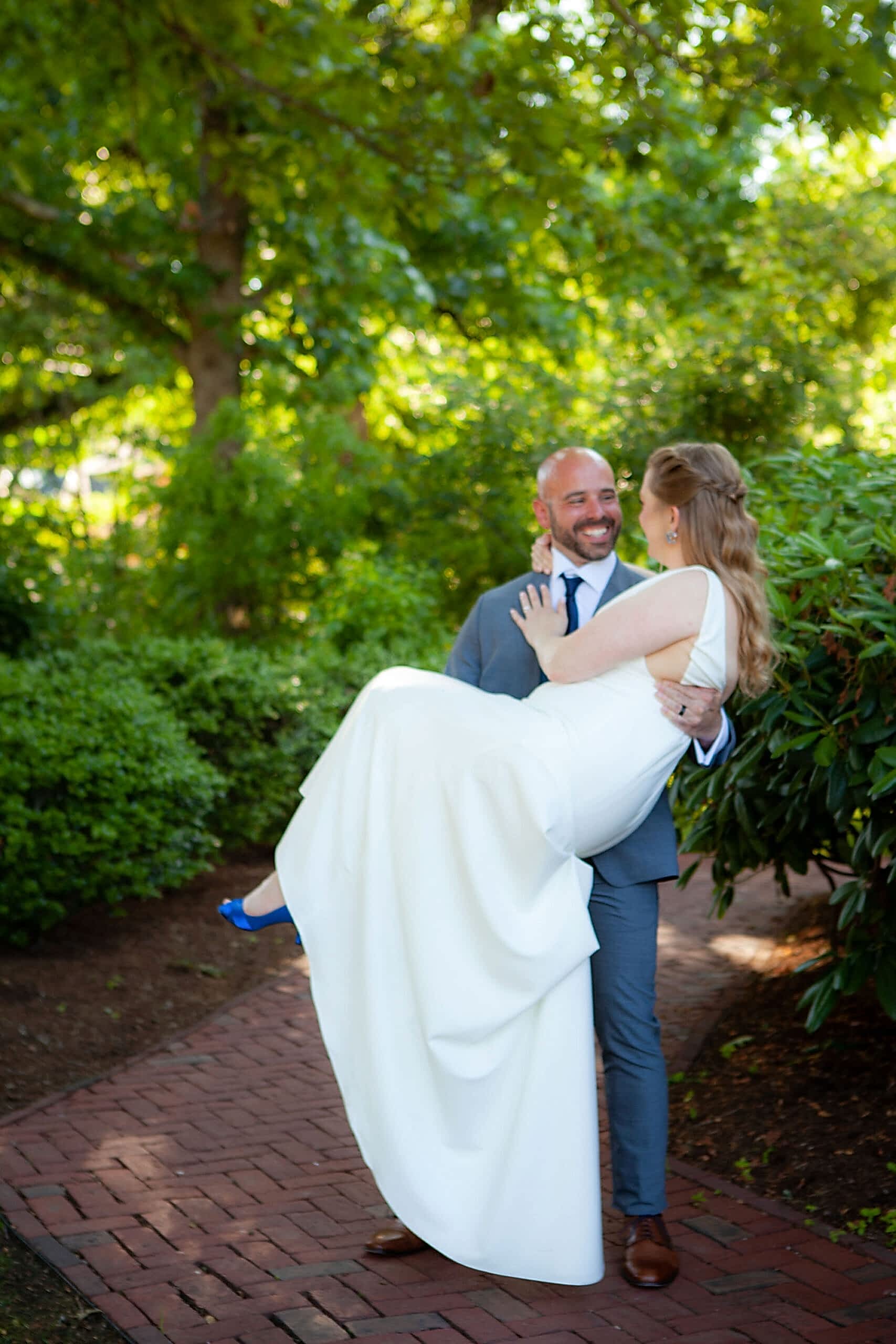 A groom in a suit carries his smiling bride in a white dress along a garden path surrounded by green trees and bushes.