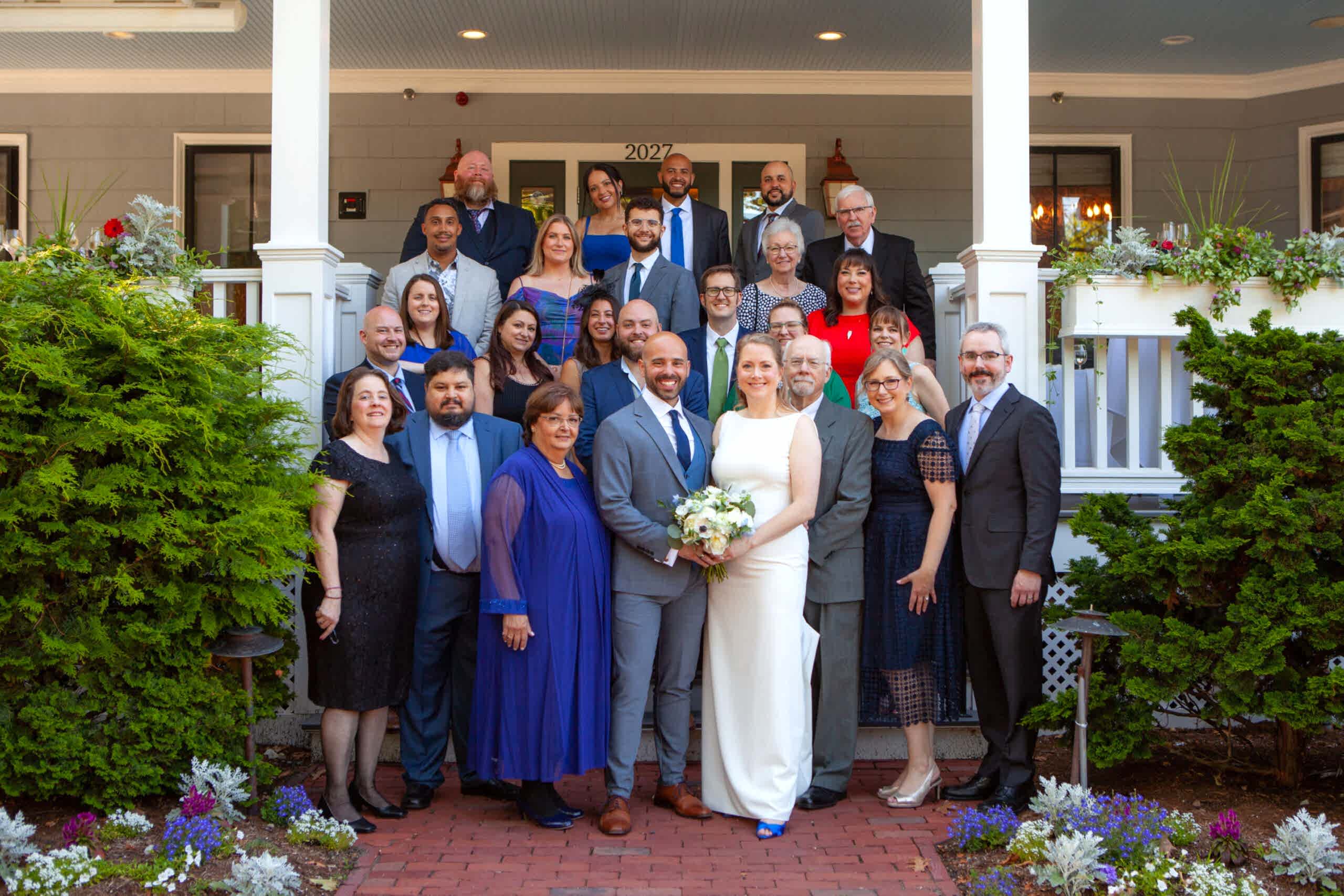 A wedding couple poses with family and friends on a porch, all smiling; the bride holds a bouquet and everyone is dressed in formal attire.