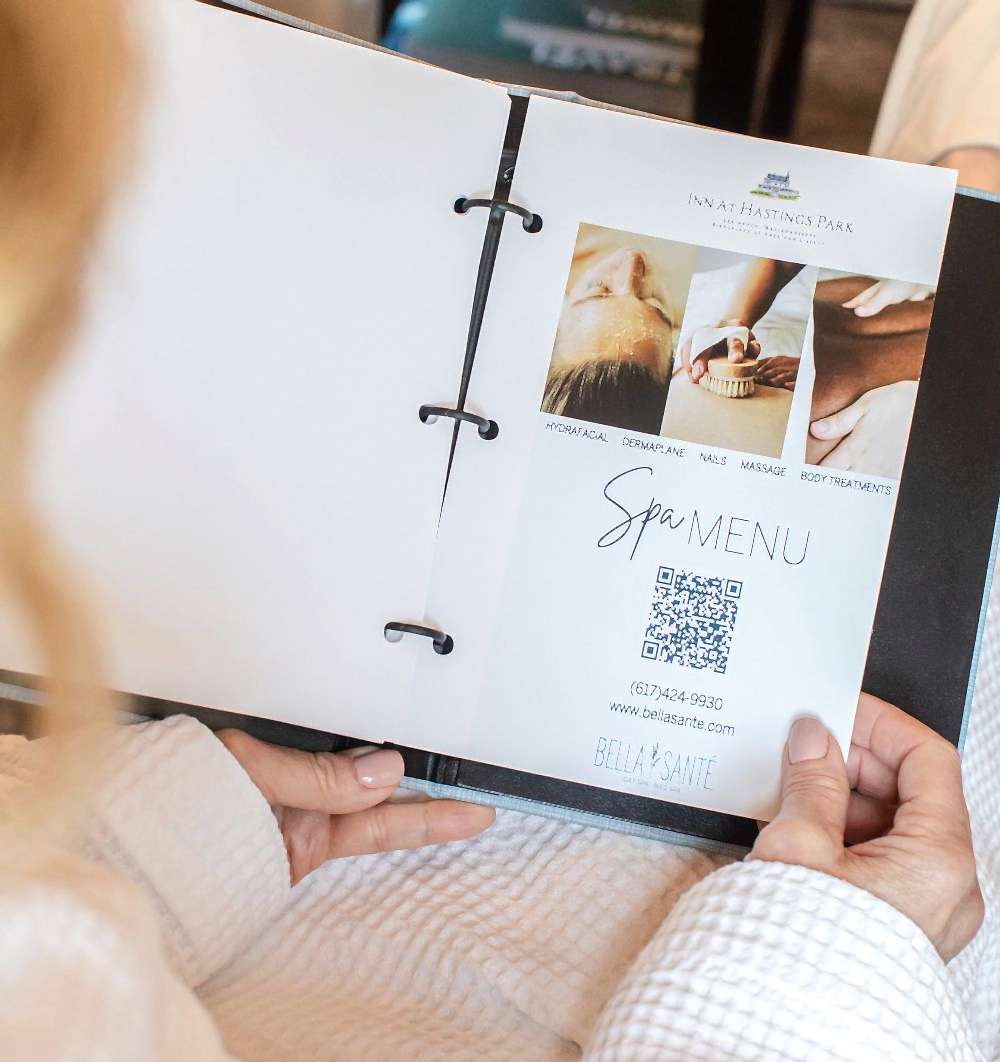 Person in a robe holding a binder open to a spa menu with images, QR code, and contact details for Bella Santé at Inn at Hastings Park.