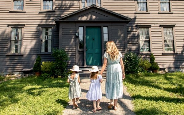 A woman and two young girls in summer dresses walk toward the green front door of a large, brown wooden house on a sunny day.
