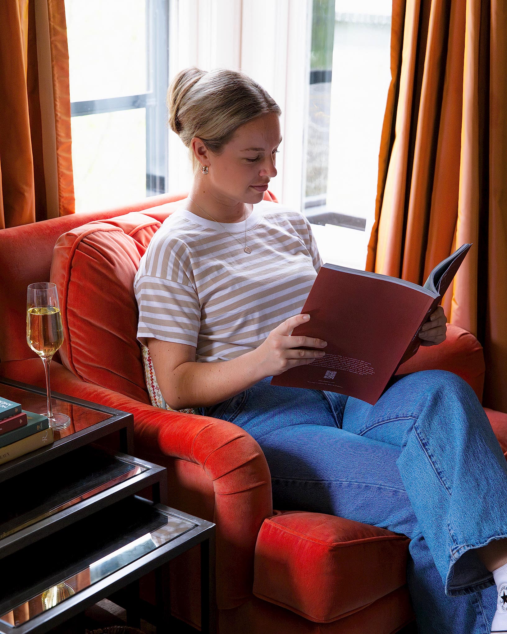 A woman sits in an orange armchair, reading a book, with a glass of champagne on a side table.