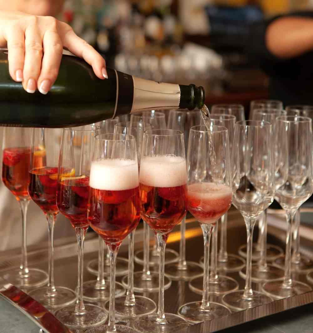 A hand pours sparkling wine into flute glasses filled with a red drink and berries, arranged on a tray next to empty glasses.