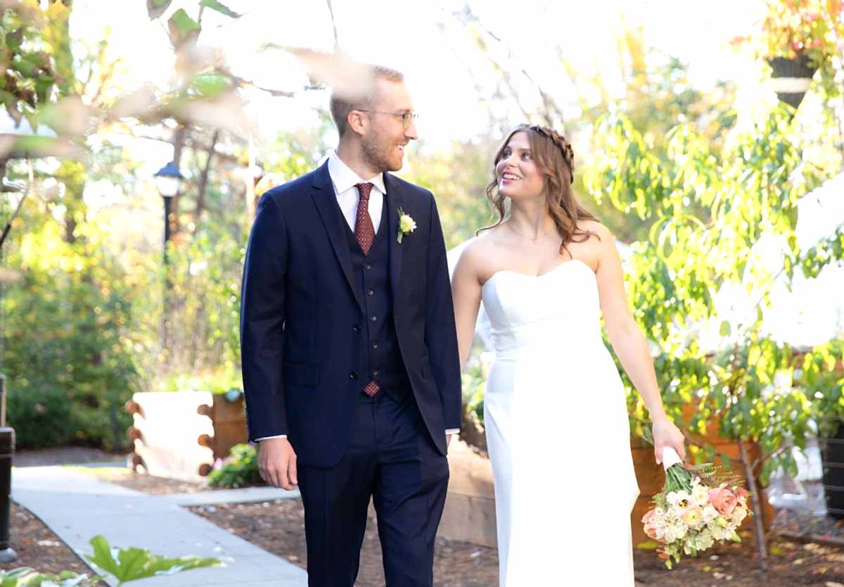 A bride in a strapless white dress and groom in a navy suit walk outside, smiling at each other, surrounded by greenery and sunlight.
