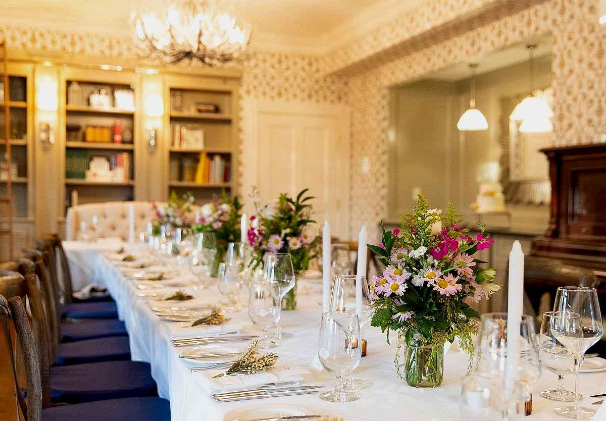 Elegant dining room with a long table set for a meal, decorated with flowers, candles, glassware, and bookshelves in the background.