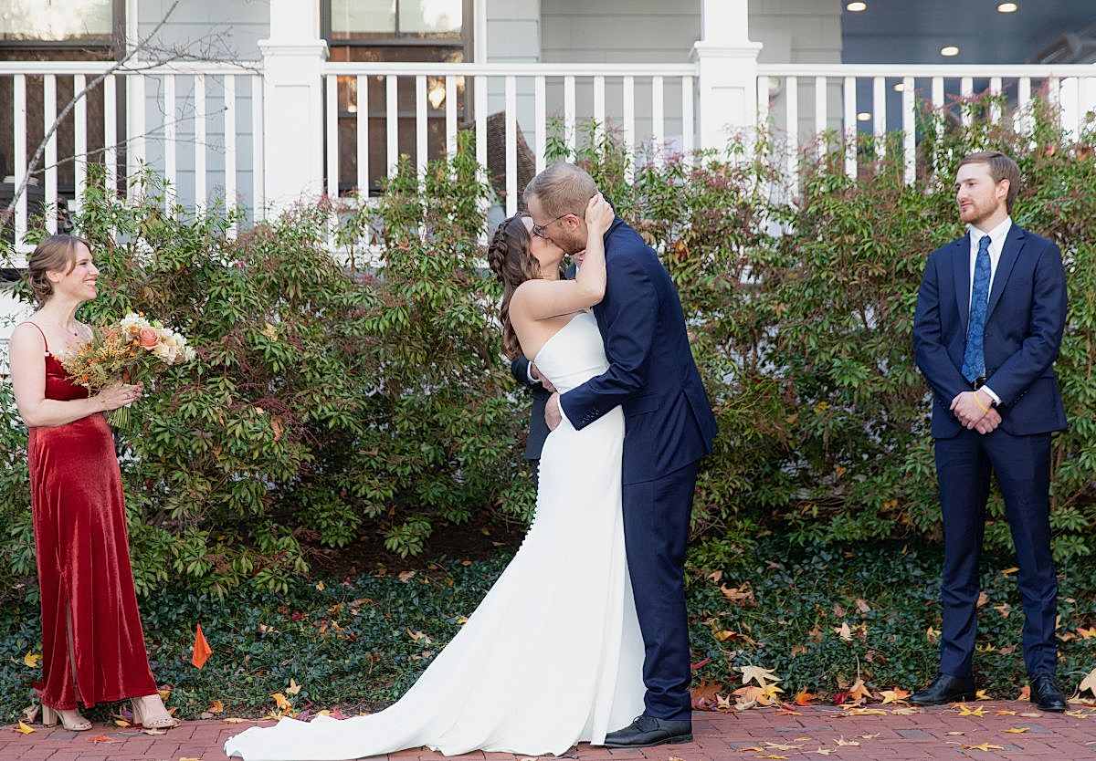 A bride and groom kiss outdoors during their wedding ceremony, with a bridesmaid holding flowers and a groomsman standing nearby.