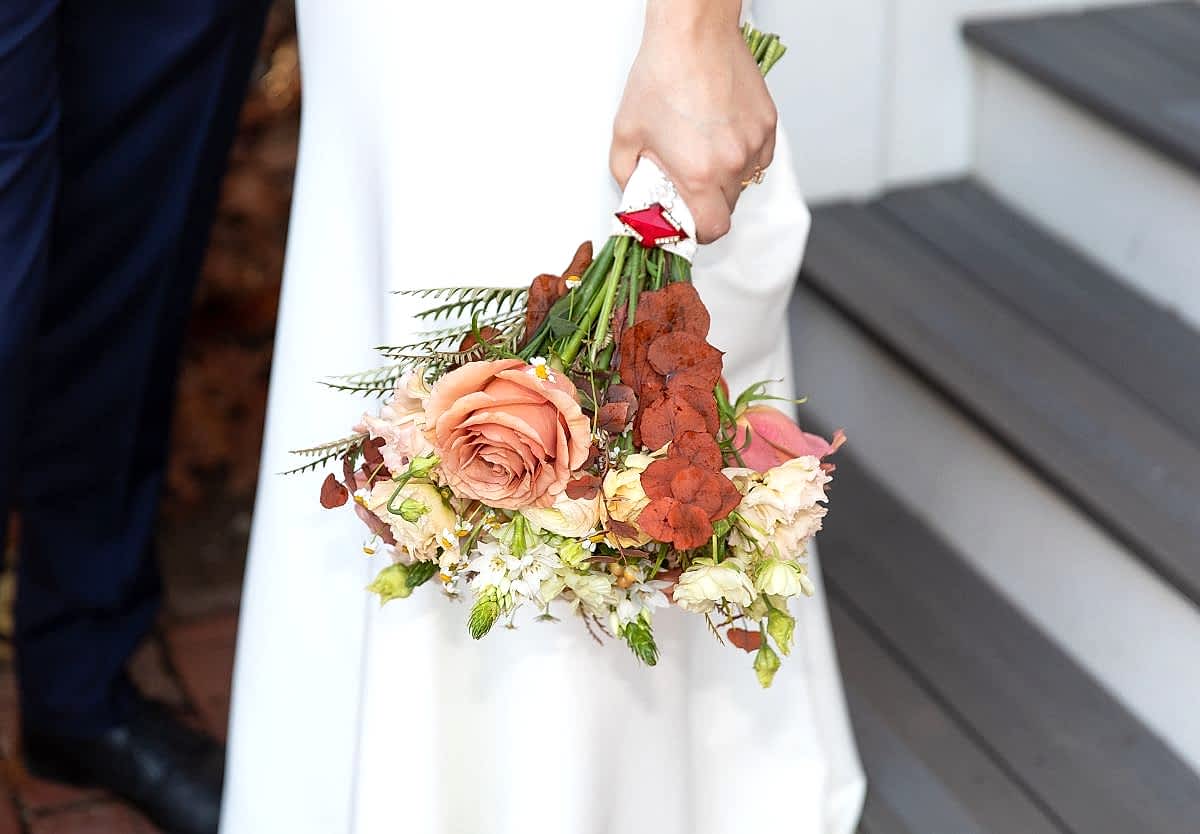 A person in a white dress holds a bouquet of mixed flowers, including a peach rose, near gray steps.