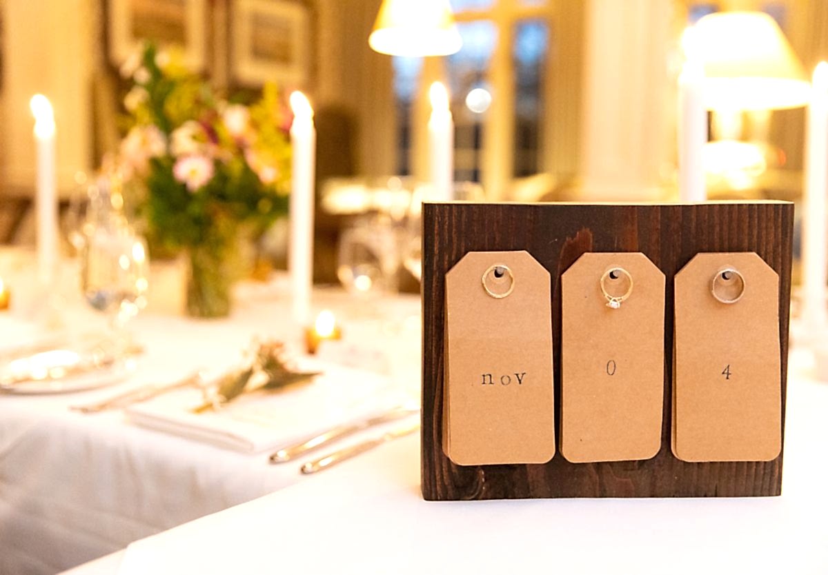 A wooden sign with tags displaying nov 04 on a table set for a formal dinner with candles, flowers, and white linens in the background.