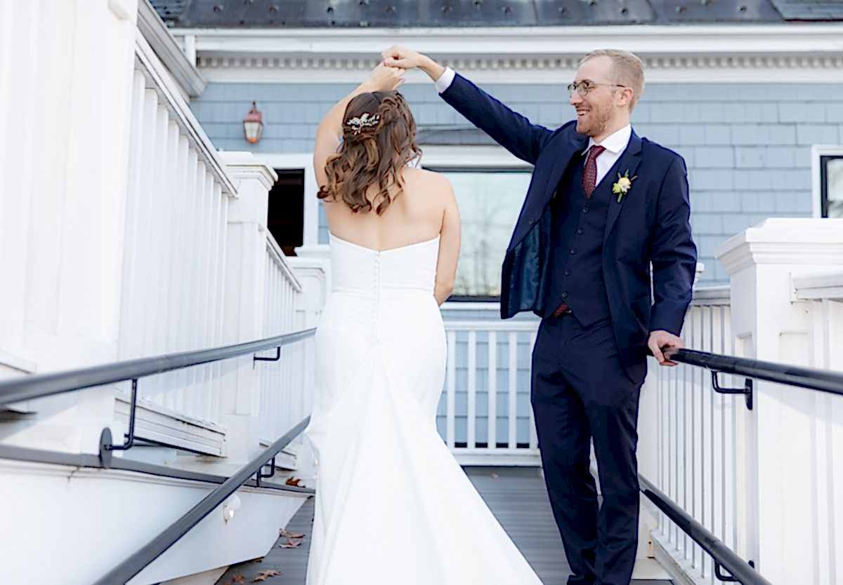 A groom in a navy suit spins his bride in a white dress outdoors on a wooden walkway, both smiling joyfully.