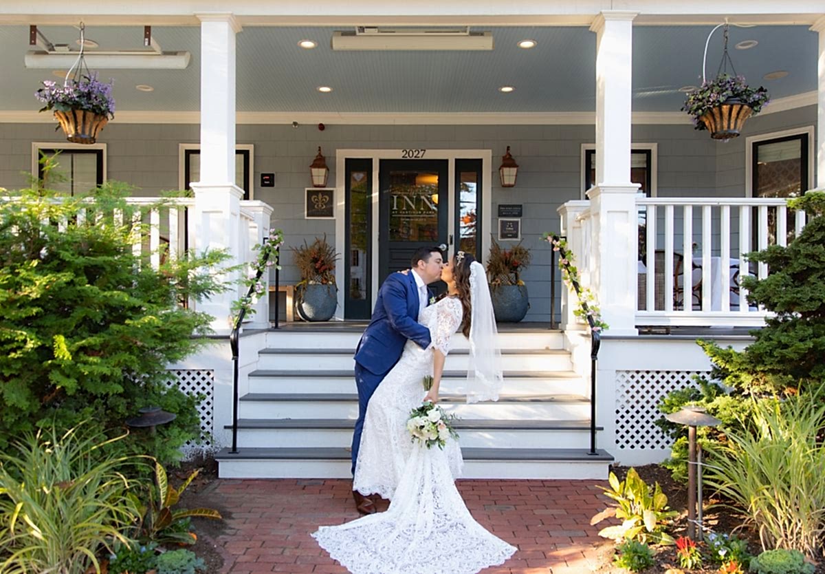 A groom in a blue suit dips and kisses his bride in a white dress on the steps of a porch decorated with flowers.