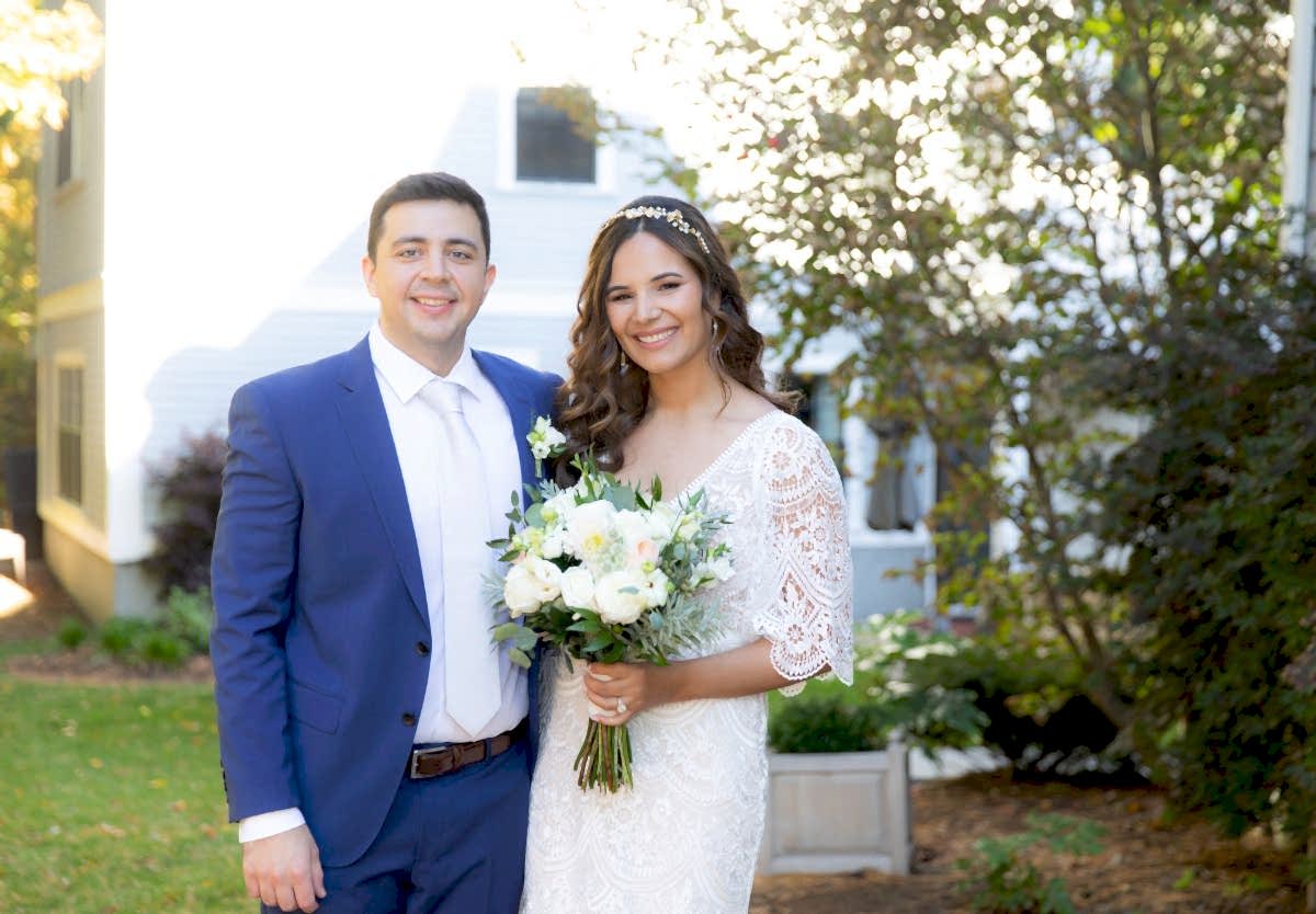 A bride in a white dress holds a bouquet, standing next to a groom in a blue suit, both smiling outdoors in a garden setting.