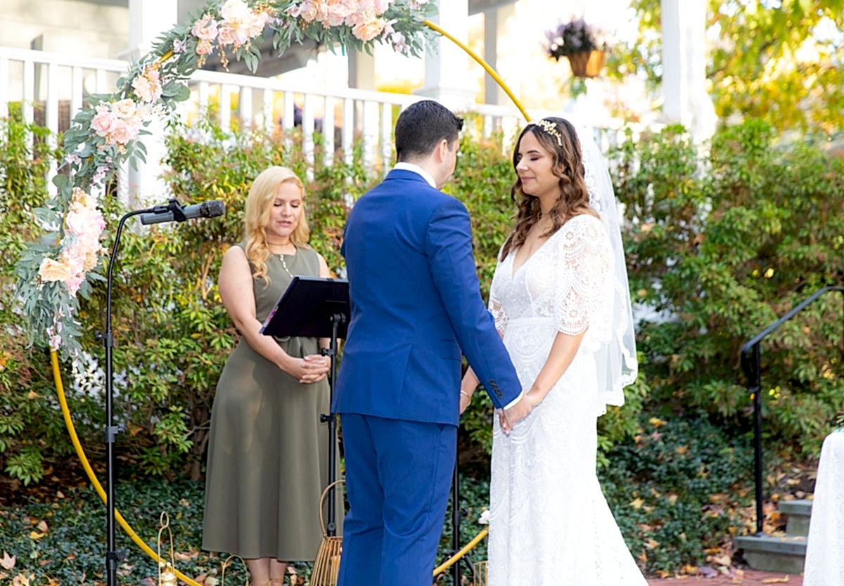A bride and groom hold hands facing each other during an outdoor wedding ceremony, with an officiant standing beside them under a floral arch.