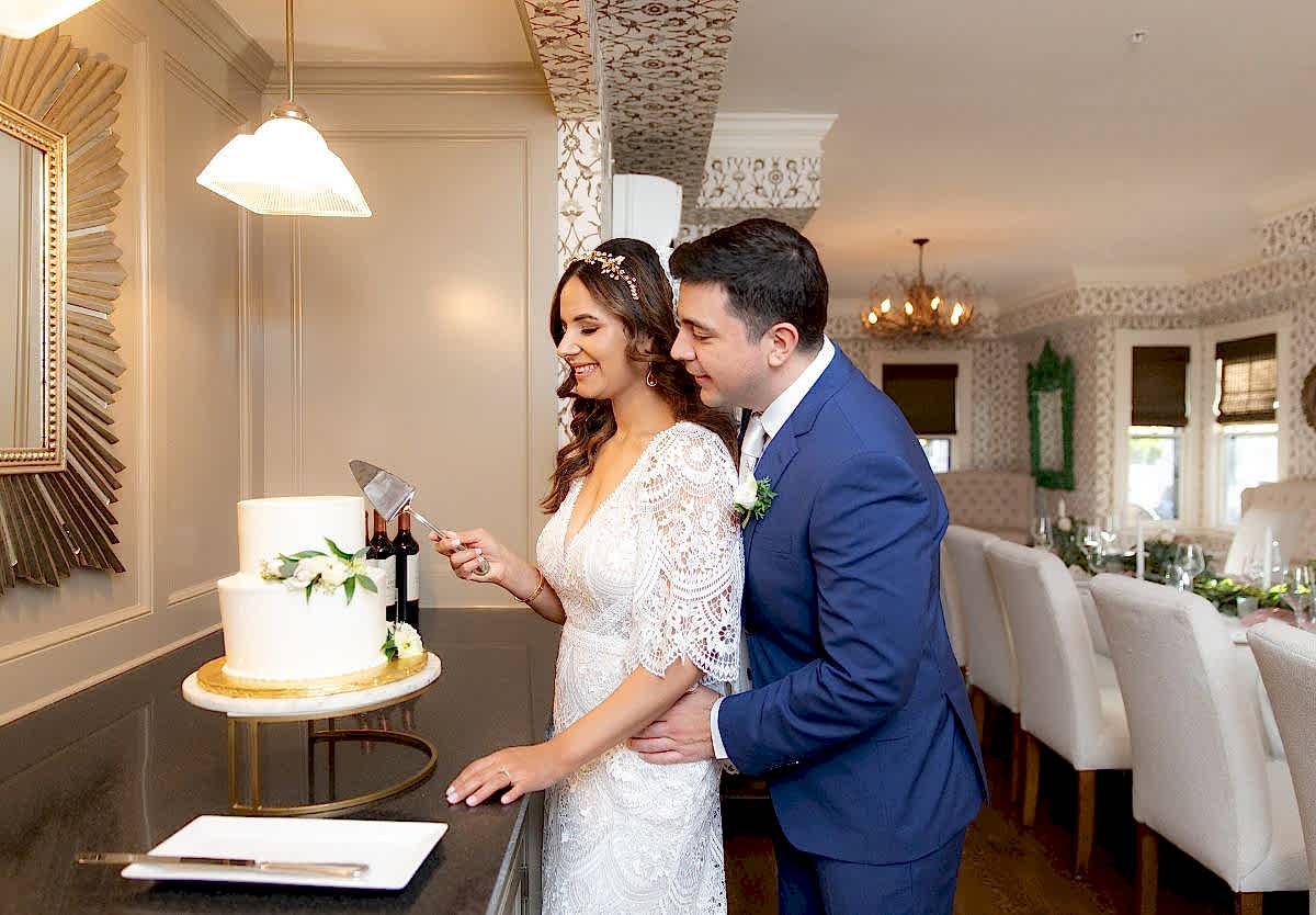 A bride and groom in wedding attire smile as they cut a white cake together in a warmly decorated room.
