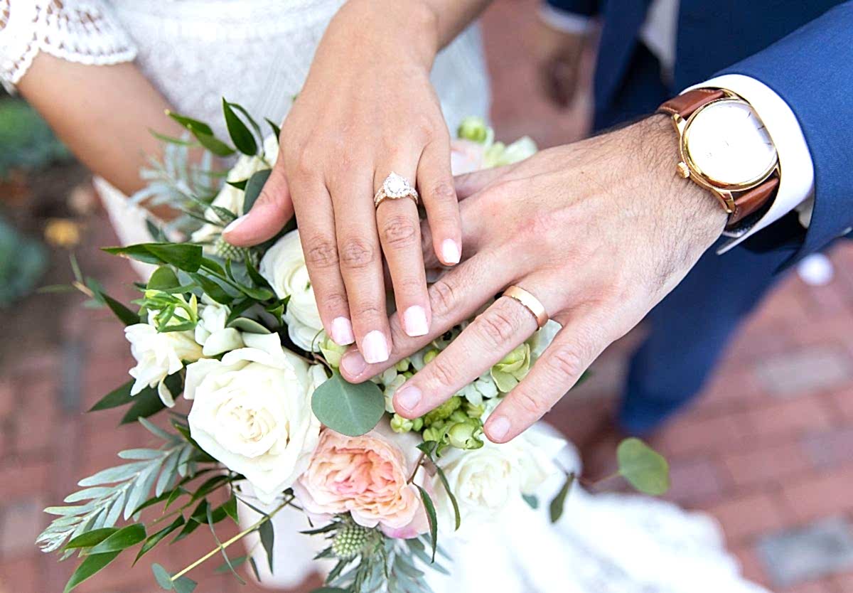 A close-up of a bride and groom’s hands with wedding rings, resting on a bouquet of white and pink flowers.