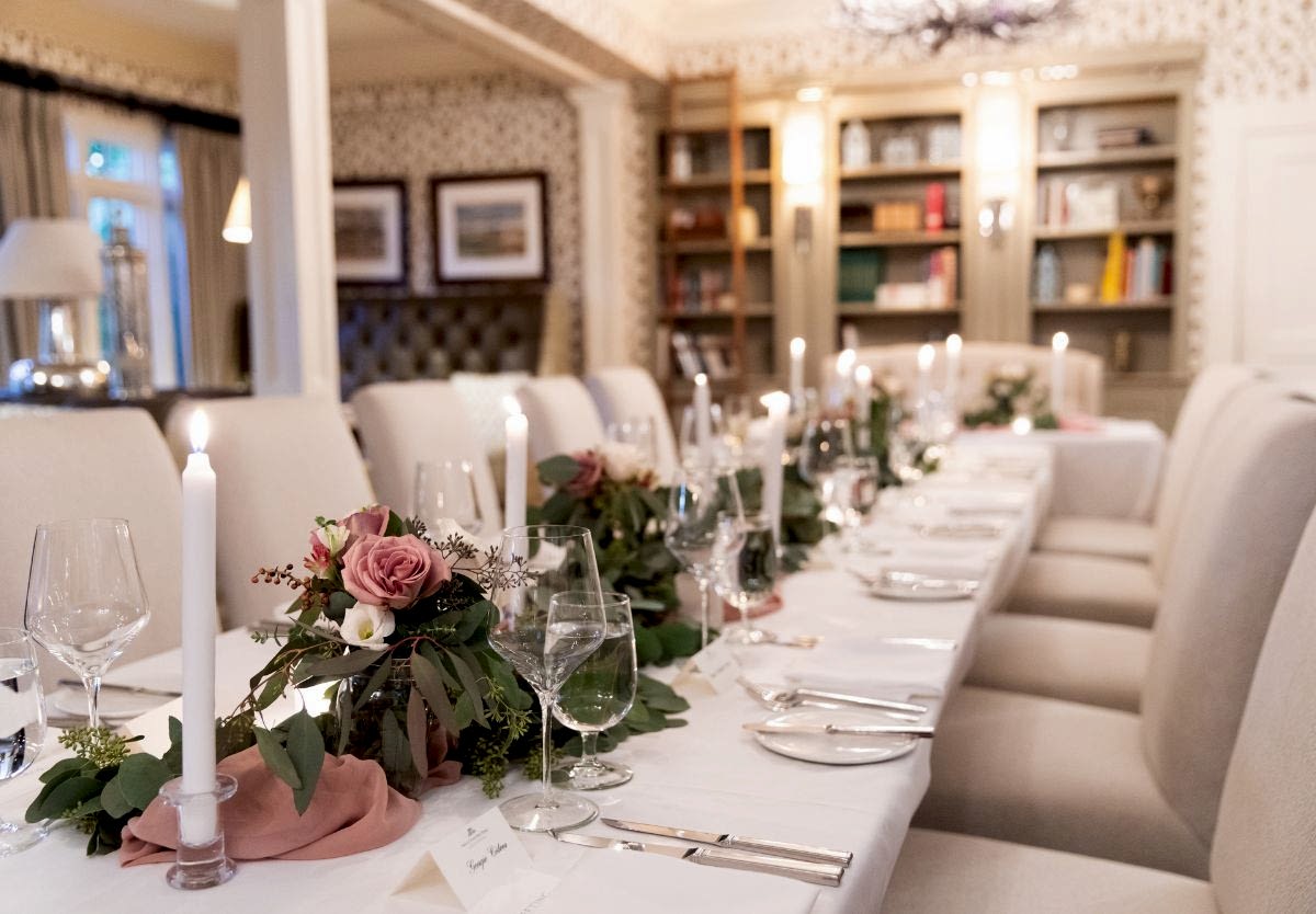 Elegant dining table set for a formal event with candles, wine glasses, and floral centerpieces in a cozy, well-lit room with bookshelves.