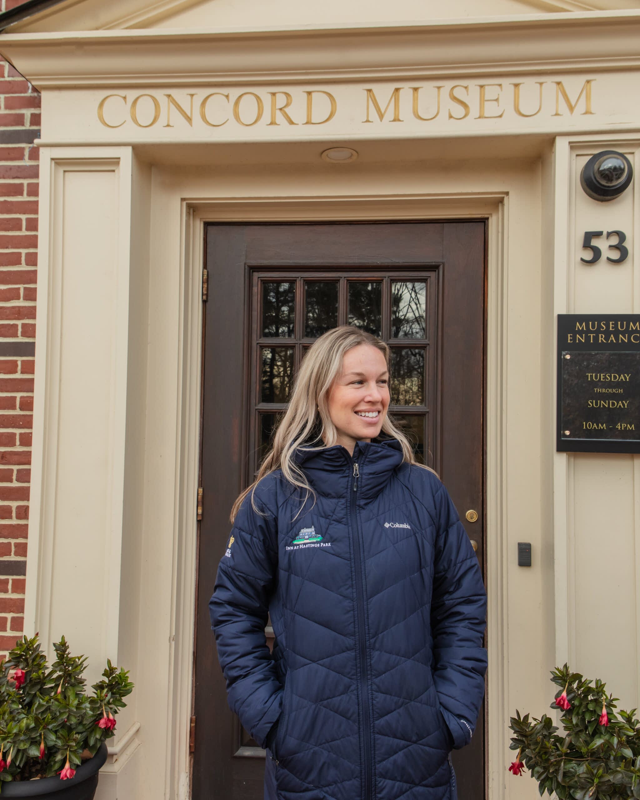 Smiling woman in a navy jacket stands in front of the Concord Museum entrance, with plants and signage visible beside the door.