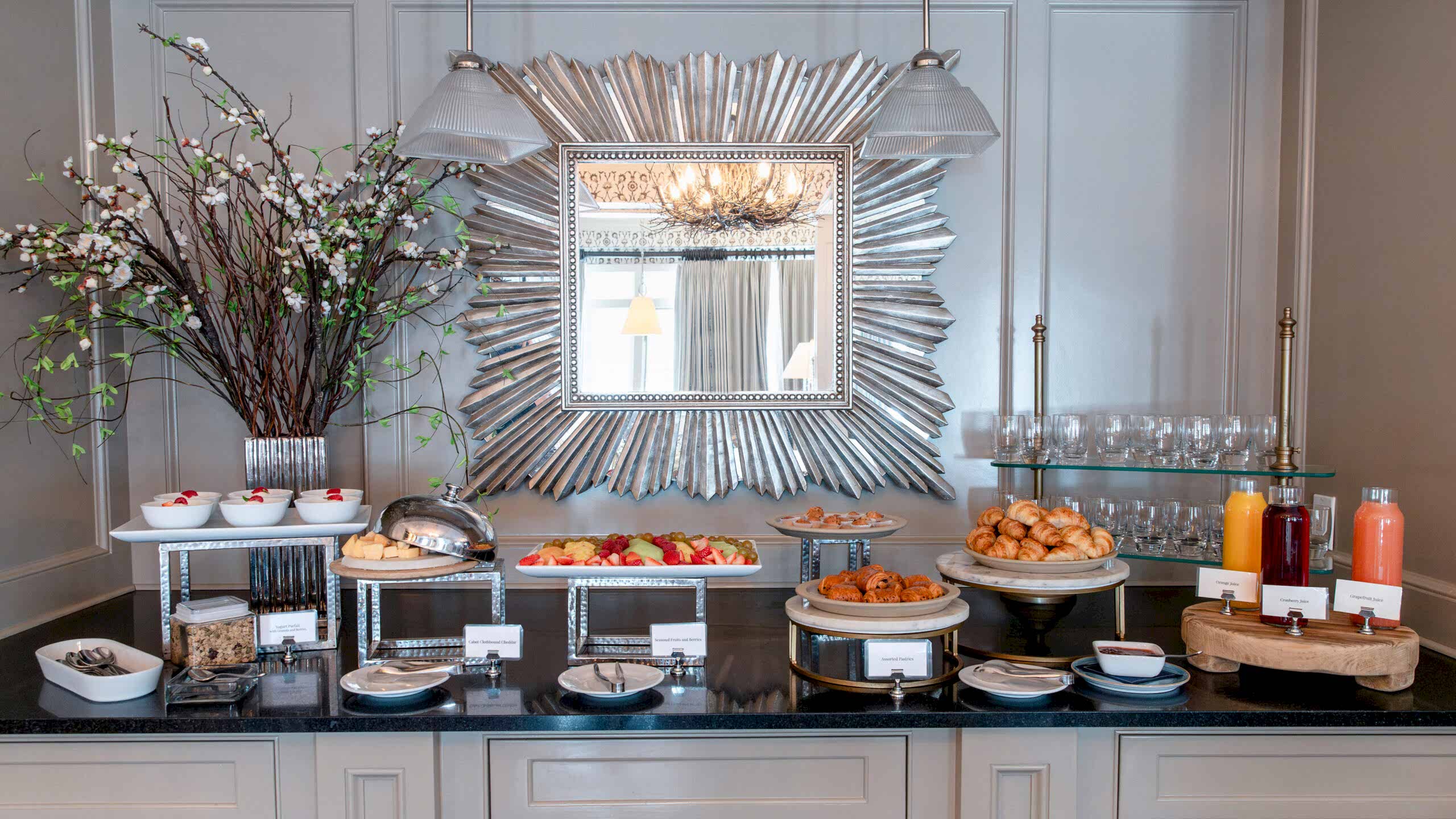 Elegant breakfast buffet with pastries, fruit, juice, yogurt, and cereal, set on a counter with a large decorative mirror and flower arrangement.