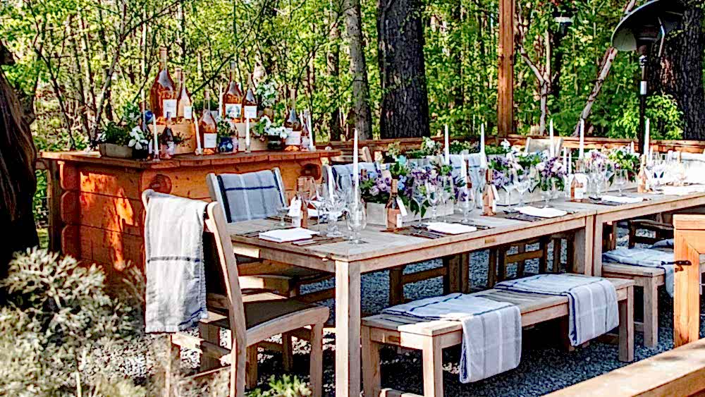 Outdoor wooden table set for a formal meal with floral centerpieces, wine bottles, and folded napkins, surrounded by trees and greenery.