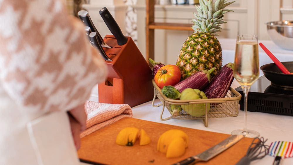 A cutting board with sliced peach, a fruit basket with pineapple and eggplants, knives in a block, and a glass of sparkling drink on a kitchen counter.