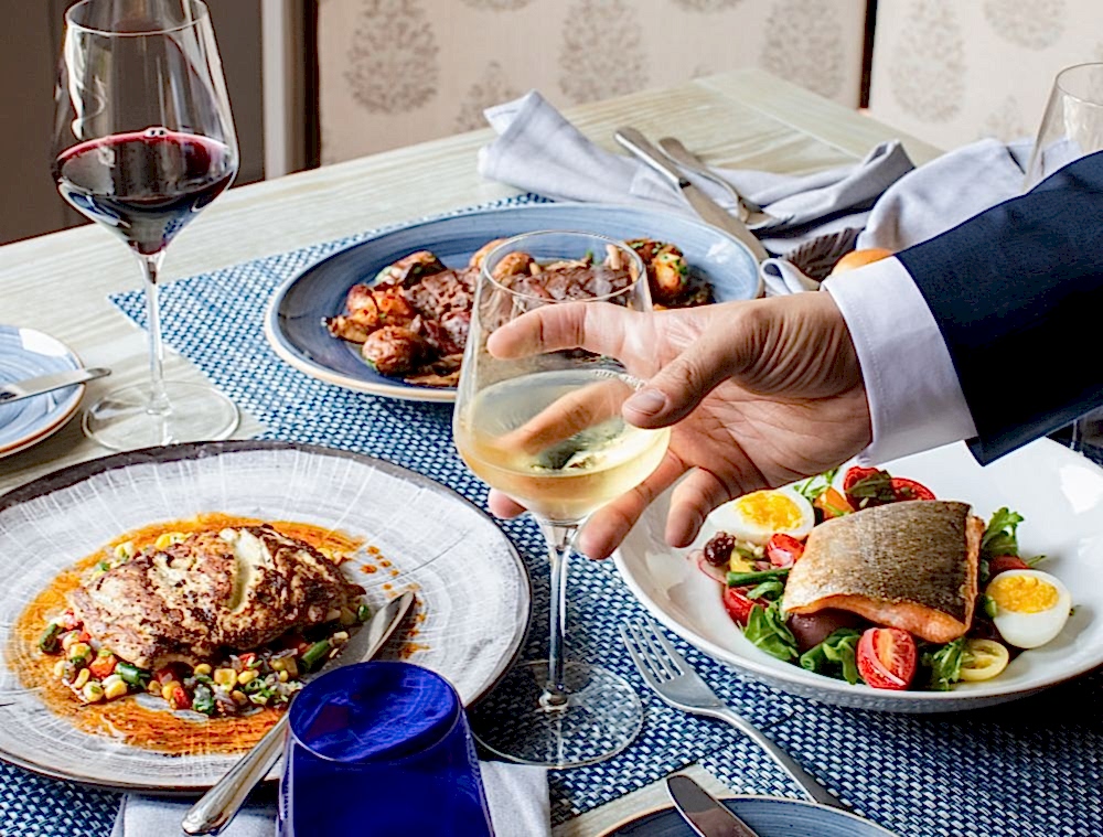 A hand holds a glass of white wine over a table set with gourmet dishes, including fish, meat, vegetables, and a glass of red wine.
