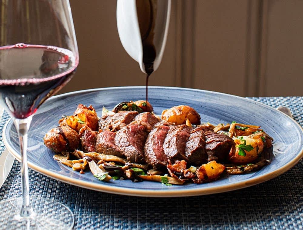 A glass of red wine beside a plate of sliced steak, roasted potatoes, and vegetables, with sauce being poured over the meal.