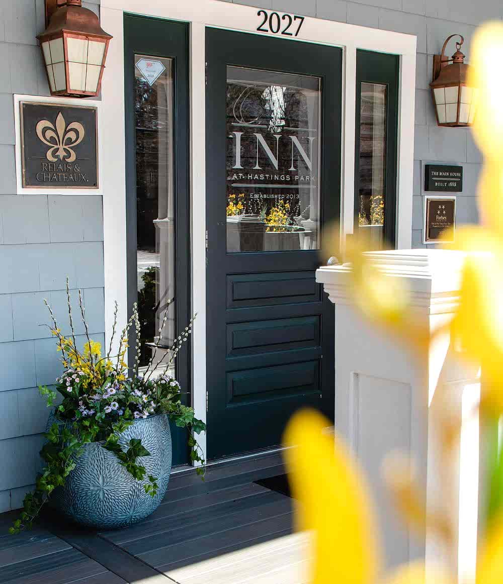 Front entrance of an inn with a dark door, potted flowers, lantern lights, and a Relais & Châteaux sign on the blue-gray wall, viewed through yellow blooms.
