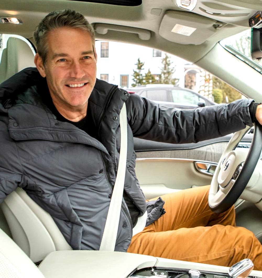 Smiling man in a dark jacket sits in the driver’s seat of a car, wearing a seatbelt, with houses and trees visible outside the window.