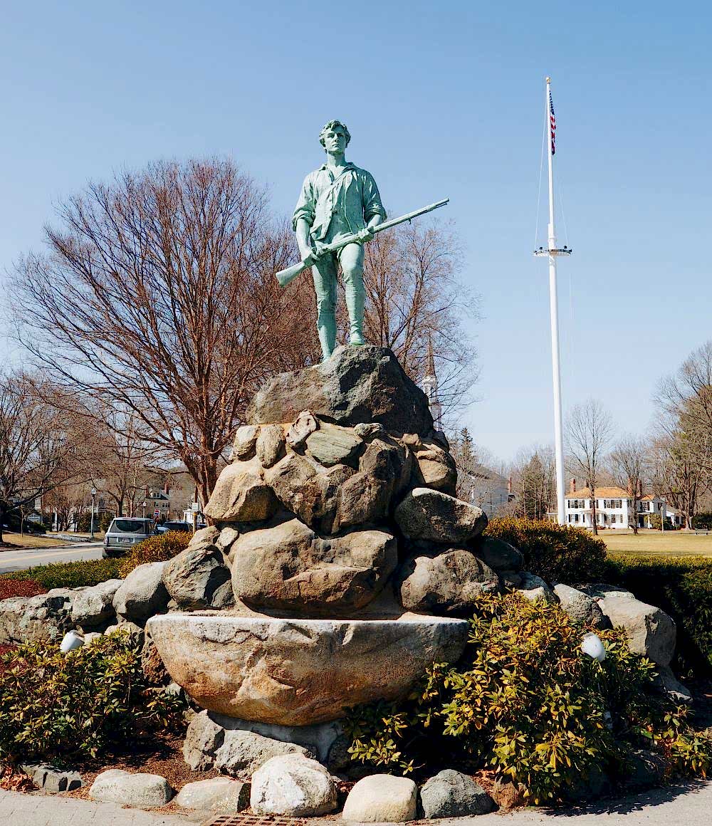 A statue of a soldier holding a rifle stands atop a rock monument, with leafless trees and an American flag in the background.