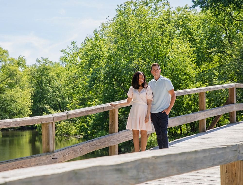 A couple stands together on a wooden bridge surrounded by lush green trees, enjoying a sunny day outdoors.