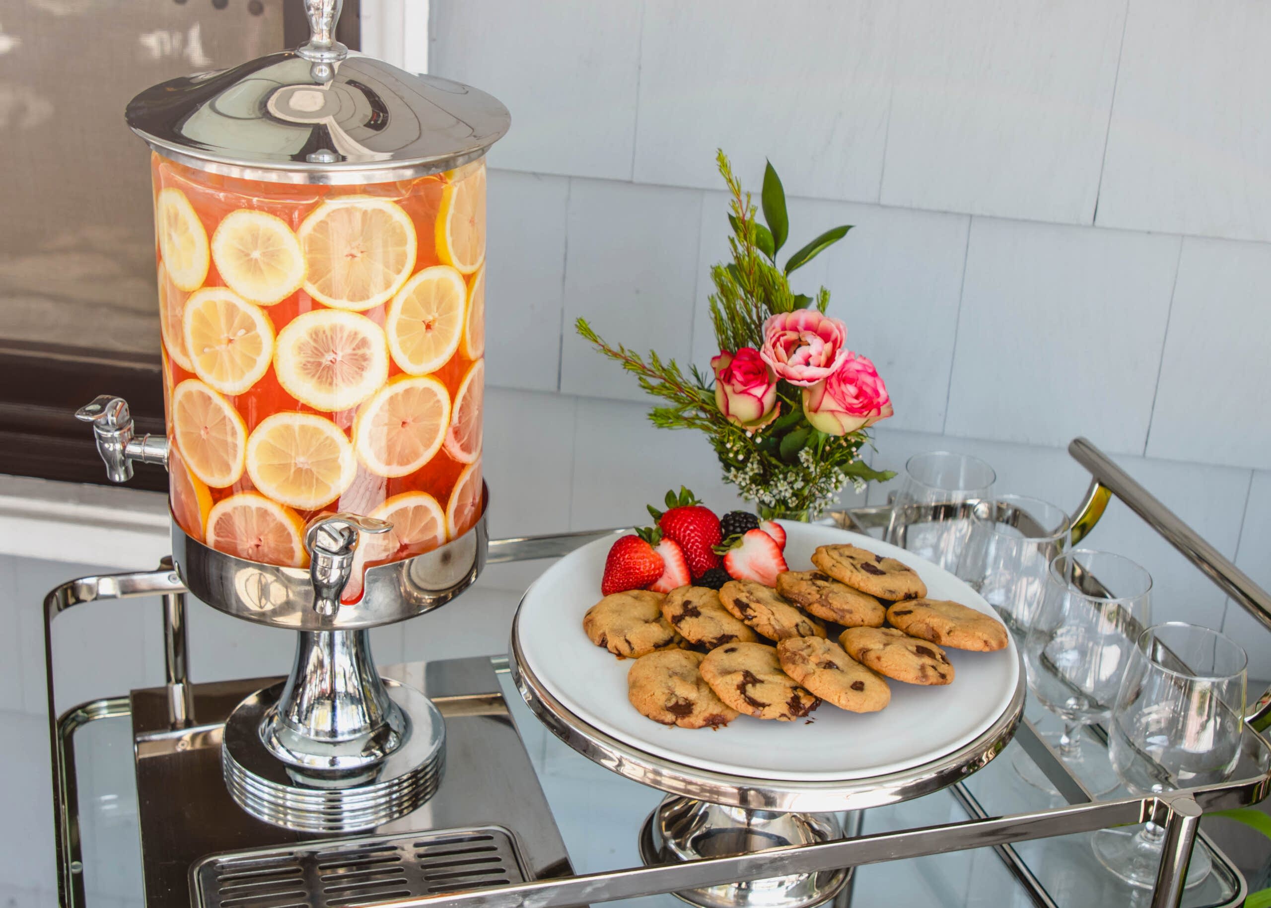 A drink dispenser with lemon slices, a plate of chocolate chip cookies, strawberries, and a small flower arrangement on a serving cart.
