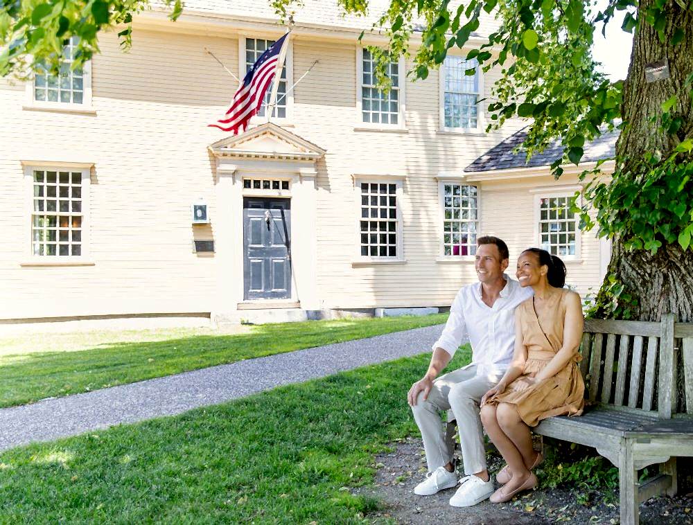 A smiling couple sits on a bench under a tree in front of a yellow house with an American flag.